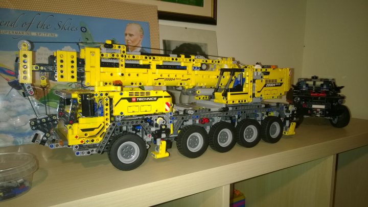 The LEPIN "LEGO" for non sensitive types - Page 84 - Scale Models - PistonHeads - The image showcases a Lego model of a yellow construction crane with a variety of parts depicted in different levels of assembly. The crane model, which is the main focus of the image, is situated on a wooden shelf. Behind the crane, there's a black construction toy, a small portion of a person visible, and a poster in the background. The space suggests a home or a workspace dedicated to collecting and assembling such toys.