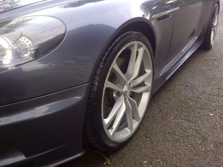 DBS wheels on DB9 - (more or less) - Page 1 - Aston Martin - PistonHeads - The image features a close-up view of the front wheel and part of the bumper of a grey luxury car, showcasing its alloy wheels with a shiny surface. The car appears to be parked on a concrete surface, and the tire also includes a silver hub cap. The car has a sleek body with a polished finish, and the image's focus and composition emphasize the details of its design and quality.