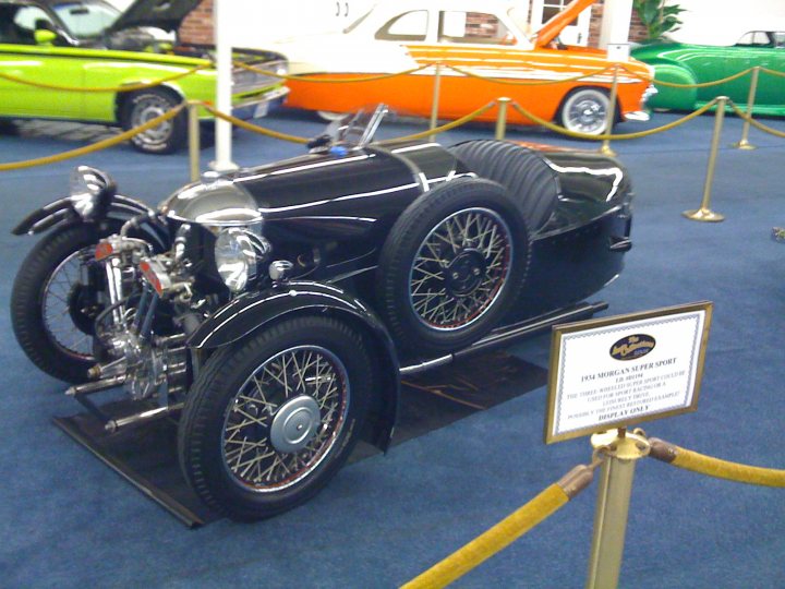 Car idents please - Page 1 - General Gassing - PistonHeads - In the heart of a car showroom, attention is captivated by a striking contrast of colors. A vintage race car, painted in pristine white, stands proudly on a black pedestal. The car is adorned with elegant copper wheels and features a vibrant red and black striped hood, adding to its classical racing aesthetics. 

Adjacent to the vintage car on the right, a 1957 Corvette display is showcased. Its lime green body is accented with white trim, representing a classic moment in automotive history. The Corvette and the race car, two vehicles from different eras, share the stage in this setting. The room they occupy is filled with other cars of varying models, indicating a diverse collection of automobiles on display.