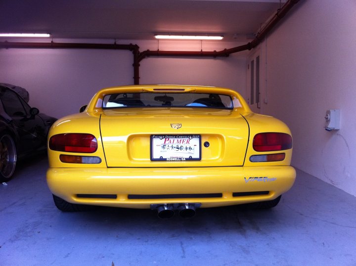 Long Pistonheads Worth Wait - The image captures a vibrant yellow sports car parked inside a spacious garage. The car, with its curved lines and round headlights, stands out against the backdrop of the garage. The garage itself is well-lit by overhead fluorescent lights, highlighting the clean and shiny surface of the car. A black car can be seen parked nearby, adding a contrast to the bright yellow of the sports car. The license plate of the sports car reads "PMP 801", indicating it's a Californian vehicle.