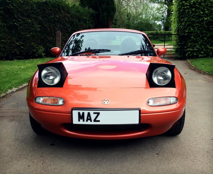 New Eunos Owner...  - Page 1 - Mazda MX5/Eunos/Miata - PistonHeads - The image shows a Mazda Miata sports car parked on a paved driveway. The car has a vibrant orange paint color and a black plastic front bumper with the word "Maz" on it. In the background, there is a line of hedges and a green lawn, suggesting a suburban or rural setting. The sky is visible above the driveway, indicating partly cloudy weather conditions. The style of the vehicle is reminiscent of sports cars from the 1980s to 1990s, with a smooth and sleek design.