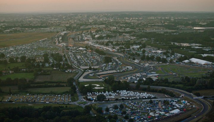 Take me back!!! - Page 1 - Le Mans - PistonHeads - This image portrays an expansive aerial view of a densely developed area at either sunrise or sunset, as indicated by the warm and golden hues illuminating the scene. The landscape is dominated by a sprawling complex of roads, buildings, and green spaces. Notably, the roads feature an interlocking network of highways and roads, weaving a complex tapestry across the area. This intricate web of roads contrasts with the mostly uniform greenery of the natural spaces. There are numerous structures, some densely packed while others are smaller and spaced out, all neatly arranged within the confines of the human-made boundaries. The topography is gently undulating, with no signs of significant elevation, giving way to the continuous expanse of the natural environment.