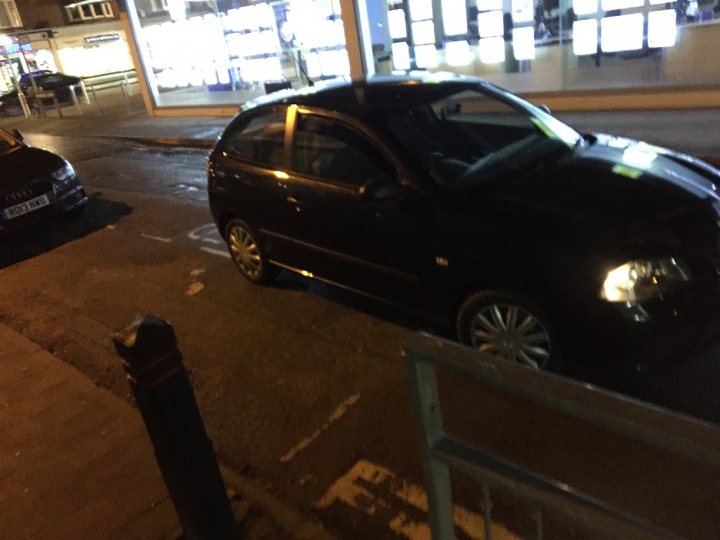 The BAD PARKING thread [vol3] - Page 498 - General Gassing - PistonHeads - This image captures a scene on a city street at night. A black Audi TT is parked on the side of the road near a car which is partially outside the frame, possibly a second Audi or a different make of car. The Audi TT has alloys and a black roof, and it is currently stationary with its headlights off, revealing a dark exterior that contrasts with the illuminated cityscape. The street appears to be wet, with visible puddles that catch the light and reflect the surrounding city structures. There are also neon lights visible in the background, indicating open businesses lining the street. The sky is clear, suggesting a cold winter night.