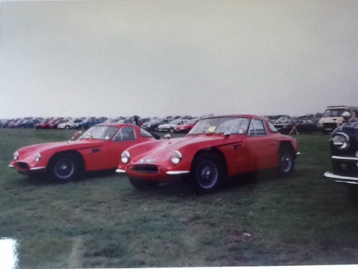 Early TVR Pictures - Page 10 - Classics - PistonHeads - The image portrays a vintage scene on a lush grass field. There are three historic sports cars parked side by side, painted in striking hues of red and white, and one is a unique mix of red and black. The cars are meticulously aligned, exuding their nostalgic charm. The backdrop reveals a row of parked cars, and a house serves as a quaint residence, adding a touch of elegance to the overall setting. This tableau transports viewers into a world of automotive history.