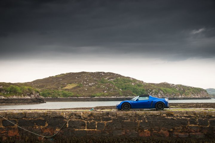 RE: Lotus Elise S2/Vauxhall VX220 | Better Together - Page 1 - General Gassing - PistonHeads - The image showcases a dramatic sky, likely indicating an impending storm. Dominating the foreground is a sleek blue sports car parked by a rocky shoreline. The vehicle's vibrant color stands out against the somber tones of the sky. In the distance, a lush green hillside stretches across the horizon, providing a stark contrast to the overcast sky. A stone wall runs along the lower part of the image, adding an element of man-made structure to this natural landscape. The overall composition suggests a moment of calm before a storm in a serene coastal location.