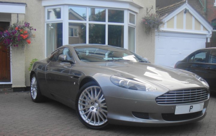 The Proper Colour for an Aston thread - silver, greys, black - Page 1 - Aston Martin - PistonHeads - The image shows a stylish gray luxury car parked in front of a house. The car is positioned at a slight angle, with the front facing to the right. It features a prominent grille with a logo at the center, which is characteristic of the Aston Martin brand. The vehicle is equipped with alloy wheels and has a sleek, streamlined design. The car is parked on a gravel driveway, which leads up to a garage. There are potted plants visible on either side of the driveway, adding a touch of greenery to the scene.