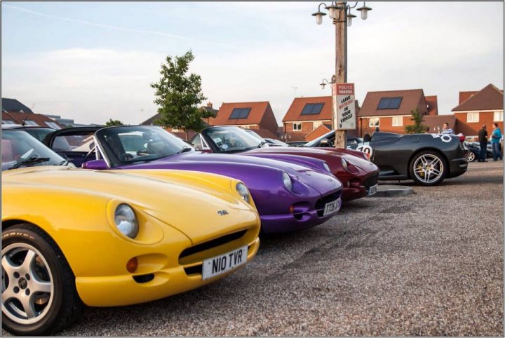 TVRCC + Guests Berkshire June Meet - Page 1 - Thames Valley & Surrey - PistonHeads - The image shows a row of classic sports cars parked on a street. They range in color, with cars visible in yellow, purple, and black. The cars are parked parallel to the curb, and there is a vintage police car stationed at one end of the row. A row of street lights lines the sidewalk nearby, and the sky is overcast. In the background, there is a building and a person standing near it. The scene suggests a relaxed atmosphere, possibly a gathering of gearheads or a classic car exhibition.