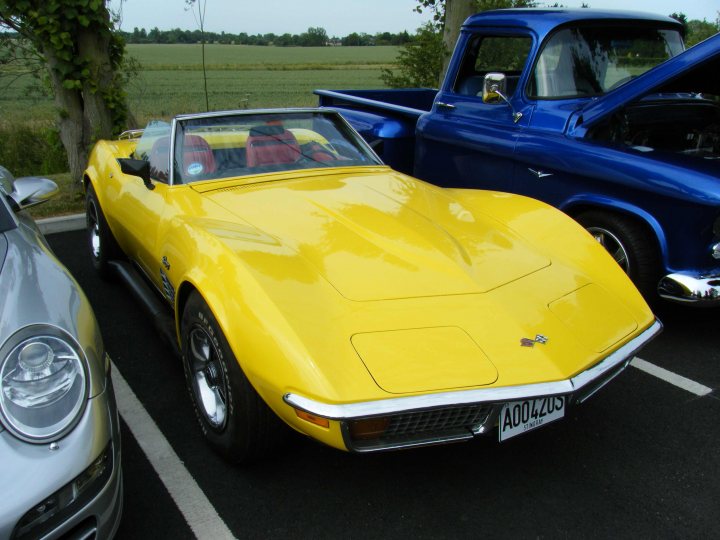 Breakfast Meeting, The Hare, Roxwell 4th June - Page 1 - Kent & Essex - PistonHeads - The image shows a vintage yellow convertible parked in a small space with a dark blue Ford truck behind it. The car's vintage design suggests it's from an earlier era, possibly the 1960s. The parking lot appears to be in a rural setting with open fields visible in the background. The reflective surfaces of the vehicles suggest that the photograph was taken in natural sunlight.