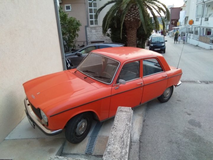 COOL CLASSIC CAR SPOTTERS POST!!! Vol 2 - Page 1 - Classic Cars and Yesterday's Heroes - PistonHeads - The image features an old orange car parked on the street next to a sidewalk. The car has a distinctive front grille and rounded headlights, typical of older vehicle models. In front of the car, there is a small concrete structure that might be used for parking markings or as a barrier. There are no people in the immediate vicinity of the car, and the background consists of a building with a palm tree on the sidewalk, suggesting a warm climate. Another car is partially visible parked further away, but it is quite small and blurred in the frame, indicating it further down the road.