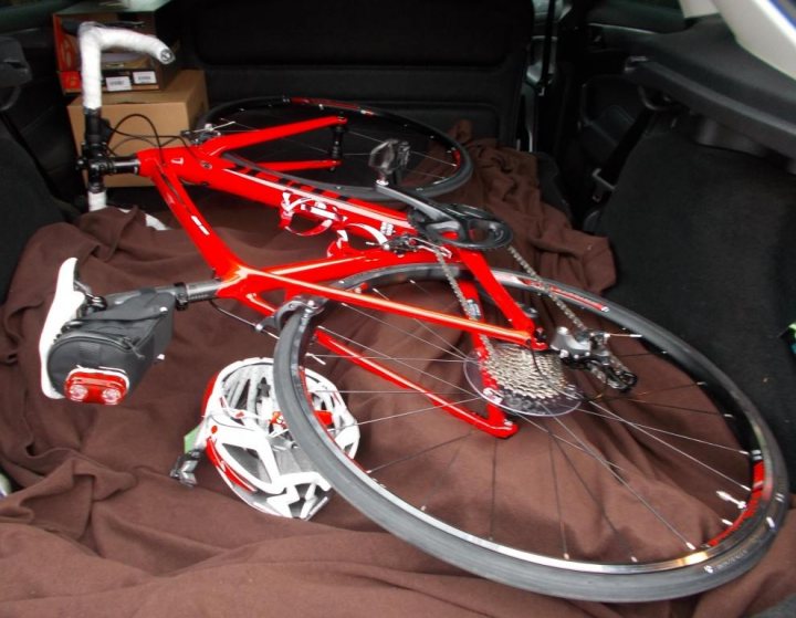 Decathlon/B'TWIN bikes (Sorry! It's a what bike?) thread... - Page 3 - Pedal Powered - PistonHeads - This image captures a scene in the trunk of a car, where a red bicycle with black tires is prominently featured. The bicycle is placed on a vibrant blue blanket, which covers the trunk floor. A black helmet, presumably for the cyclist, is positioned next to the bicycle. On the left side of the image, there's a small, light-colored step stool, suggesting that it might have been used to lift the bicycle into the trunk. The interior of the car is visible in the background, providing context to the location of the items in the foreground.