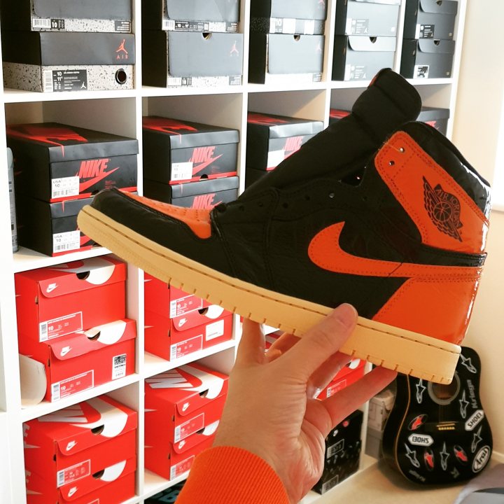 Anyone into trainers/sneakers? (Vol. 2) - Page 222 - The Lounge - PistonHeads - The image shows a person holding up a pair of Nike sneakers. The shoes are predominantly black and orange with the famous 'Swoosh' logo on them. The soles of the shoes appear to be white and brown, suggesting they might have a gum or caramel sole. The person is standing in front of a wall that has multiple shelves with various items, including what seems to be a collection of sneakers and other merchandise. The sneakers are well-lit, emphasizing their design details.