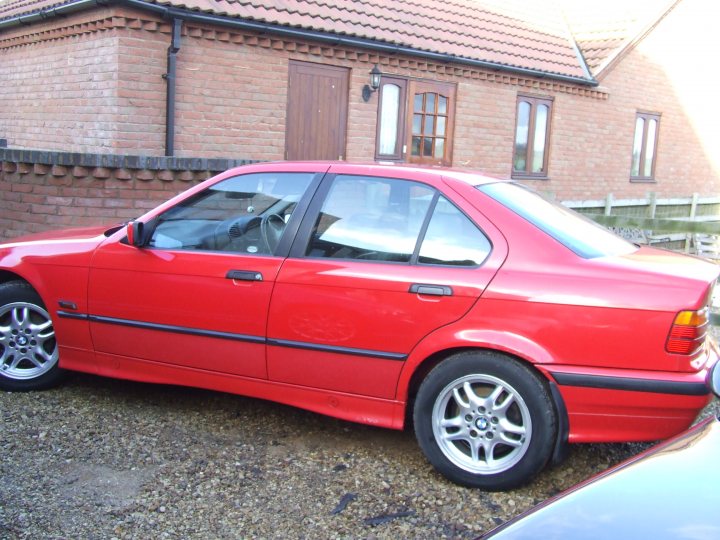 Winter Pistonheads Project - The image displays a vibrant red sedan parked next to a one-story brick building during the day. The car appears to be in good condition, with shiny wheels and a sleek design. The ground beneath the vehicle is noticeably dirty, indicating a less manicured outdoor setting. The building, which could be a garage or house, has a traditional brick exterior. There are no visible texts or distinct features in the immediate surroundings that reveal the location or context of the scene. The image is a standard, day-to-day photograph with a clear focus on the car and building.