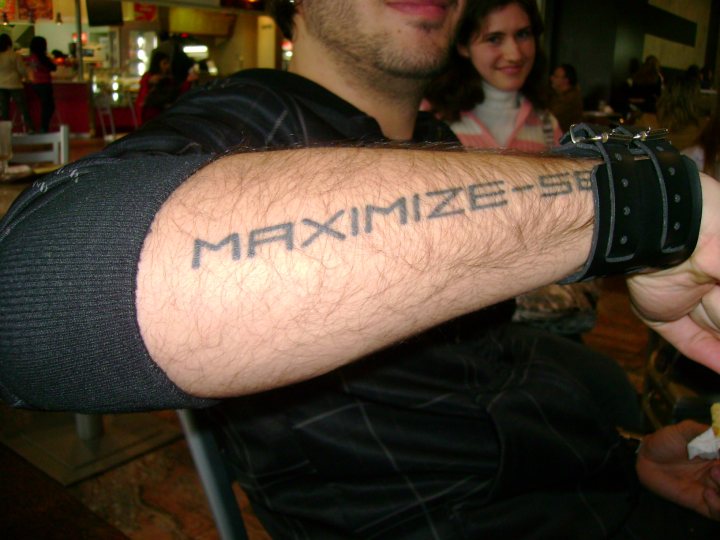 Max - In the image, a man is seated at a dining table wearing a black tank top and a wristband on his left wrist. His right arm is stretched out across the top of the table. On this extended arm, a noticeable tattoo of the word "maximize" is visible. The arm also features a black band around the upper bicep area. The setting appears to be a restaurant or dining establishment, as suggested by the presence of multiple chairs, dining tables, and other patrons in the background.