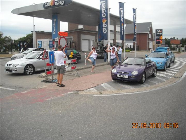 Pistonheads - The image depicts a bustling scene at a road check or toll plaza. A silver car is parked near a man standing by a traffic sign, who appears to be directing or observing the vehicles. Several people, including a woman with a camera, are walking near the parked cars, likely pedestrians in the area. Multiple cars are visible, both parked and in motion, on the road where various lanes are visible. The road is adorned with a mix of signage, including blue and white banners and roadside paraphernalia. The overall atmosphere suggests a regulated transport area with staff and vehicles navigating the area smoothly.