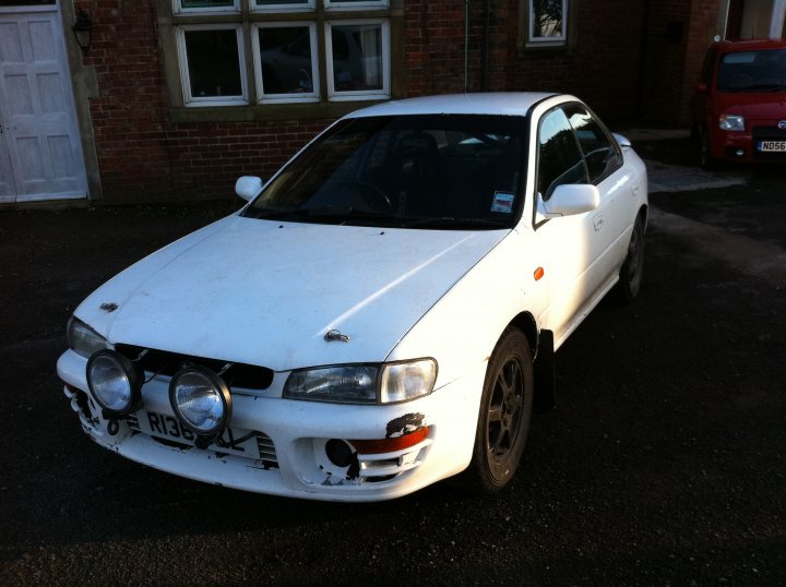 Impreza Rally Pistonheads Road Subaru - The image shows a white, four-door sedan parked on a driveway in front of a brick building. The car is positioned at an angle, and the background features a red vehicle in the distance. The vehicle in the foreground has aftermarket fog lights installed at the front. The photo is taken during the day under clear skies.