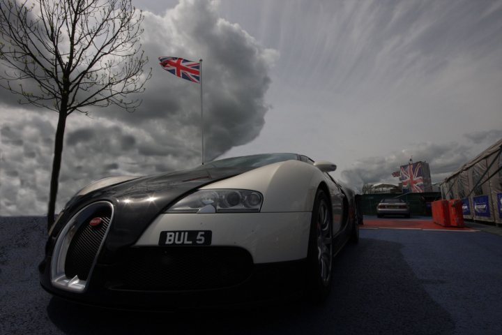 Needed Sat Pistonheads Silverstone - This image showcases a striking scene featuring a vintage white Bugatti sports car. The car, adorned with a license plate reading "BUL5," is parked on a runway painted in a rich shade of blue. Behind the sleek Bugatti, the backdrop is dominated by a cloudy sky, and boldly standing out against it, is the British flag. Adding to the intrigue of the scene is a white building with a blue roof visible in the distance. The overall composition of the image is a blend of modern elegance and patriotic symbolism.