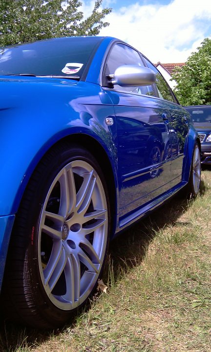 Cars On The Green - Photo Thread - Page 1 - East Anglia - PistonHeads - The image features a close-up of a silver-rimmed, blue sports car parked on grass. The car is positioned at a slight angle to the viewer, showcasing its glossy finish and the distinctive Audi emblem in front. The setting suggests a gentle breeze is in motion as suggested by the slight shadow cast by the car. The background reveals a tree-lined street, hinting at an suburban or residential area.