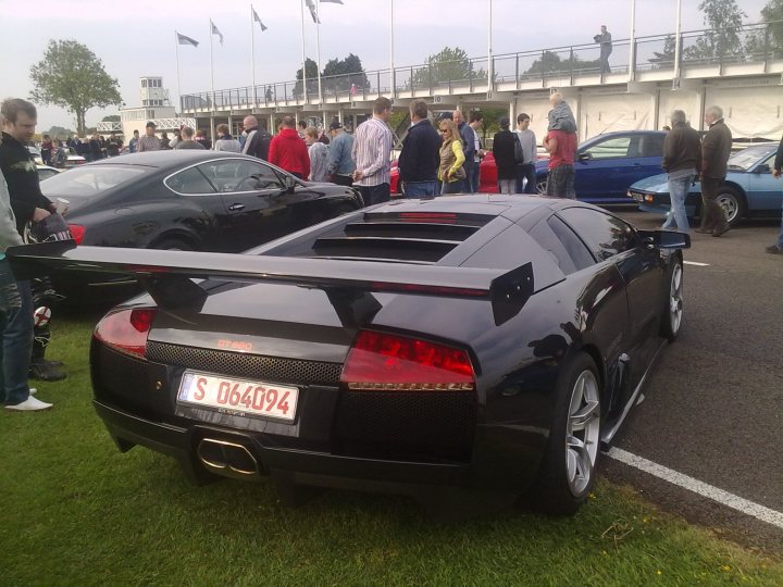 Supercar Sunday Pistonheads - The image showcases a black sports car prominently parked on a grassy plot, ready for acceleration. The vehicle is adorned with a red tail light, which stands out against its black exterior. The car is positioned near a white line, suggesting it is park ed in a designated area, perhaps at a parking lot or a racing event. In the background, various other cars are parked, conveying an impression of a crowded parking space. Among them, some people and buildings are visible, adding context to the scene.