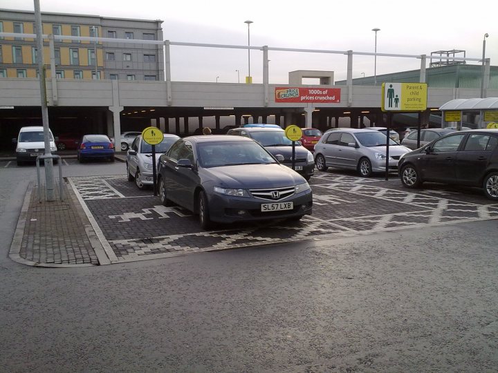 The BAD PARKING thread - Page 231 - General Gassing - PistonHeads - The image shows a parking lot with multiple cars parked. The cars vary in sizes and colors, and are parked in designated spaces. A sign with a male figure peering into a car is visible to the right, suggesting a cautionary notice for men to look for children or other people in their vehicle. The setting appears to be a busy urban area, indicated by a shopping mall in the background. The ground appears to be wet, suggesting it may have rained recently.