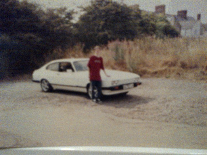 Wong's car history.  - Page 1 - Readers' Cars - PistonHeads - In the image, a young man stands in front of an old, white car. The car, a model with a prominent spoiler, is parked on a gravel road. The surroundings suggest a rural or semi-rural setting, with hedges and trees visible in the background. The man, wearing a red shirt, is positioned to the left of the car, with his gaze directed towards the vehicle. The overall atmosphere of the photo evokes a sense of nostalgia and adventure.