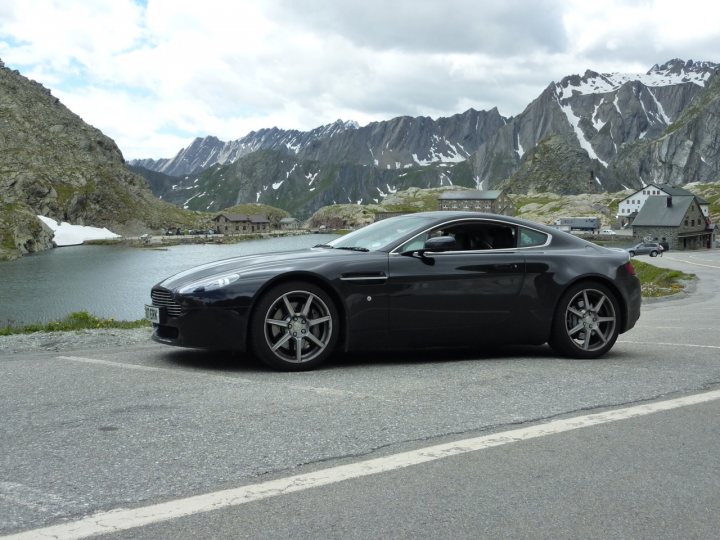 Trip of a Lifetime (picture heavy) - Page 1 - Aston Martin - PistonHeads - The image showcases a striking black Aston Martin car parked on a curvy road by a serene lake. The car's sleek design contrasts with the rugged backdrop, which features snow-covered mountains and a quaint village nestled in the valley. The mountains rise majestically into the sky, partially obscured by layers of clouds. The lake, calm and peaceful, suggests a tranquil getaway or journey for the car's driver. The overall scene exudes a sense of tranquility and adventure, appealing to both nature lovers and car enthusiasts.