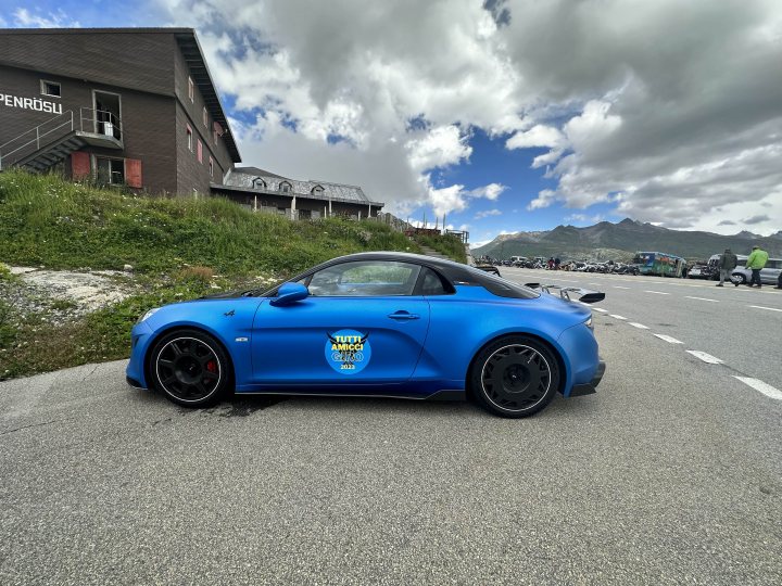 A110R “Radical” - Page 16 - Alpine - PistonHeads UK - The image shows a striking blue sports car parked on the side of a road. The vehicle is sleek, with a low-slung design and prominent wheel arches. It's positioned at an angle that allows for a clear view of its front and rear. The background reveals a mountainous landscape with a hint of snow on the peaks and a small building partially visible. There are no people or other vehicles immediately around the car, emphasizing its prominence in the scene.