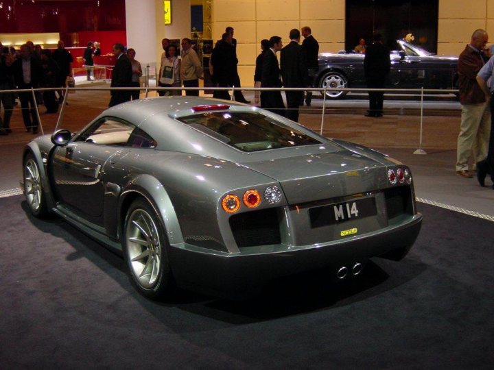 Pistonheads - In the image, a unique silver sports car with a black license plate is the center of attention, displayed in an indoor showroom. The car's sleek design and shiny exterior reflect on the glossy black floor underneath it. Beyond the car, it's a bustling scene with people milling around, possibly admiring other vehicles or engaging in conversation. The environment suggests a major car show or exhibition.