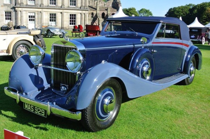 A vintage truck is parked in a field - Pistonheads - The image features a classic blue convertible parked on grass, as part of a car show. The vintage car is adorned with chrome accents and has a distinctive red stripe on its side. The car's color contrasts beautifully with the surrounding lush green grass. In the background, the entrance to an impressive brick building can be seen, suggesting that the car show is held in front of an urban or upscale setting. A couple of people are present in the area, likely attendees or organizers of the event, who add a human element to the scene. The overall atmosphere of the image is nostalgic and elegant, evoking a sense of timeless beauty.