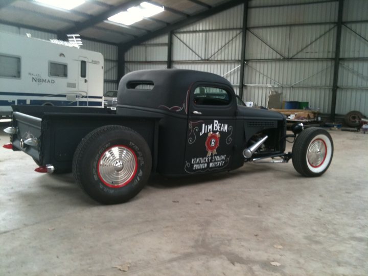 Pistonheads Ratrod Paint - The image depicts a dark-colored antique truck with a large cab and a flatbed trailer. The truck is stationed inside a large metal shed with an open side, allowing a clear view of the vehicle. The truck is branded with the words "Jim Bean" and "Kentucky Straight Bourbon Whiskey," indicating its possible use as a promotional vehicle. A classic white camper is visible in the background, suggesting a nostalgic setting.