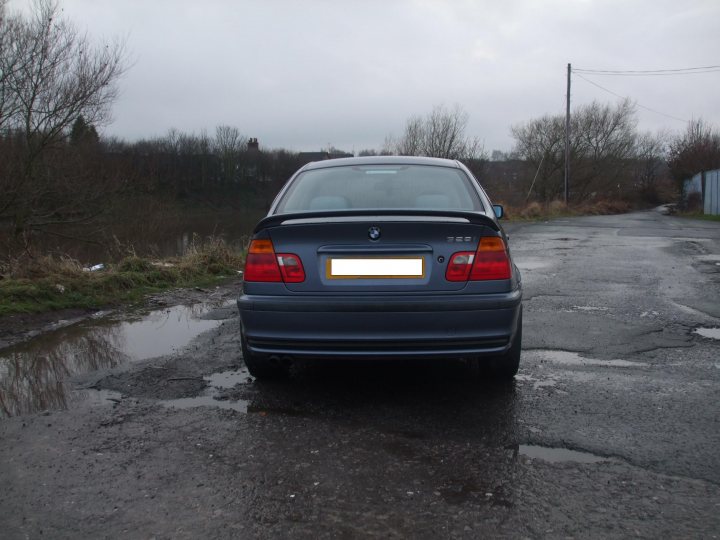 Pistonheads - This image depicts a light gray sedan parked on the side of a road that appears to be wet, suggesting recent rain. The car has a European-style license plate, and the environment around it includes barren trees on both sides of the road, implying a deserted or remote setting. There are no people visible in the scene, and the overall atmosphere is one of tranquility and solitude.