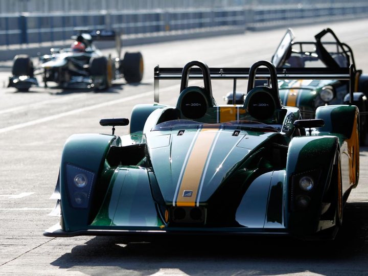 My Caterham R500 with the Caterham F1 team in Jerez - Page 1 - Readers' Cars - PistonHeads - The image showcases a dynamic scene on a race track. Two sports cars are prominently displayed, one predominantly black with gold and white stripes, and the other a striking green with a white and yellow stripe running down the middle. Both cars are equipped with open-roof designs, hinting at a convertible or exotic racing car. The cars appear to be in motion, adding a sense of speed and intensity to the image. The backdrop of the image, a tunnel through which the cars are driving, provides a sense of depth and perspective.