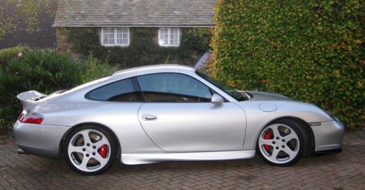 Ruf Pistonheads Lust - The image depicts a silver Porsche car parked on a brick pavement. The car is facing to the left, positioned against a backdrop of a house with a lush green garden. The car is well-maintained, gleaming under the light, with visible taillights, a sunroof, and alloy wheels with a red accent. The overall scene is serene, with the car as the central focus of the photograph.