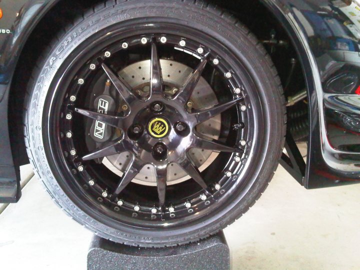 Finally finished the new wheels - Page 1 - Noble - PistonHeads - This is a close-up image showing the wheel of a car, featuring a black and silver alloy wheel with multiple spokes. The wheel is bolted to the hub through a series of lug nuts at the base, where the wheel also connects to the vehicle's suspension. The hub itself is not visible, and the tire appears to be in good condition without any notable signs of damage or wear. The car is parked on a flat surface with no sign of motion, as indicated by the absence of dynamic reflections or indicators of traction in the vinyl ground mat that can be seen below the tire. The focus is clearly on the wheel and hub, providing detail of their design and workmanship.