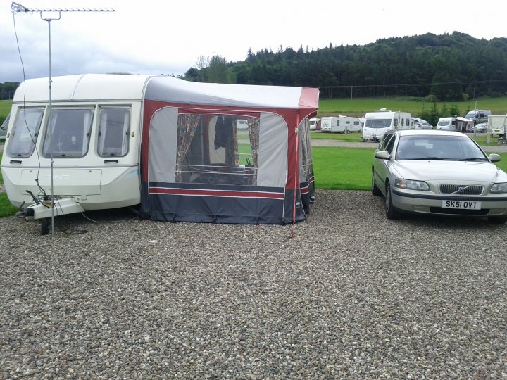 Useable caravan for £500? - Page 1 - Tents, Caravans & Motorhomes - PistonHeads - The image shows a camping site in a gravel area. A white car is parked to the right of a vintage caravanning caravan. The caravanning caravan has a matching red awning, creating a tent-like structure over its rear area. A separate tent with a grey canopy is also set up, providing additional shade for campers. There are other display trailers visible in the background, suggesting the site is a retail or exhibition area.