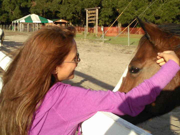 A woman petting a horse in a corral - The image captures a serene moment at a stable. A young woman with long hair is seen reaching out to pet the nose of a brown horse. She stands on a dirt path next to a fence that seems to be in a racing or training area. Behind her, a building can be seen, suggesting this could be a working farm or equestrian center. The atmosphere is tranquil, with no other people visible in the image.