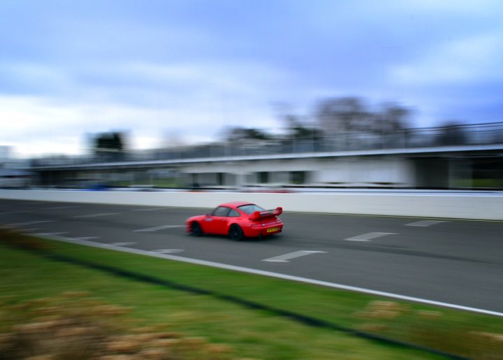 Robert Barrie Track day 24th Feb - photos - Page 1 - Goodwood Events - PistonHeads - The image captures a vibrant scene on a racetrack. A red Porsche sports car is in motion, speeding through the racetrack's infield, which is neatly maintained with grass. The vehicle's large rear wing, a distinctive feature, adds to the sporty ambiance. The track itself is racing green and appears to be empty apart from the sports car, suggesting that it might be a lone practice session or early morning run. Beyond the track, a blurred backdrop of trees can be seen, reinforcing the sense of a rural or semi-rural location for the racetrack.