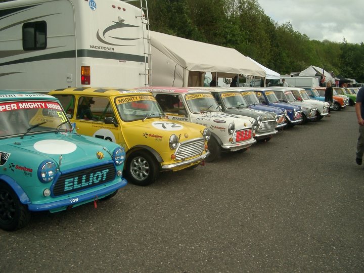 This weekend (10-11th May) - Brands Hatch - Page 1 - Classic Minis - PistonHeads - The image features a line of miniature cars parked outside. The cars are miniature replicas of famous cars and are on display at a car show. Each car is painted in a bright color and has decals and numbers on it, representing various racing teams. They are arranged neatly in a single row, parked on a gravel surface next to a recreational vehicle. The atmosphere suggests a fun and festive event.