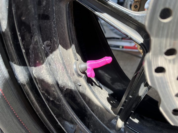 the bb trackday thread.   - Page 477 - Biker Banter - PistonHeads UK - The image features a close-up view of a vehicle's wheel and tire. There is a prominent pink object, which appears to be a toy car or some other small item, placed in the middle of the wheel. The tire has visible tread patterns and a metal hubcap is attached to it. In the background, there are blurred elements that suggest an industrial setting with machinery and tools present.