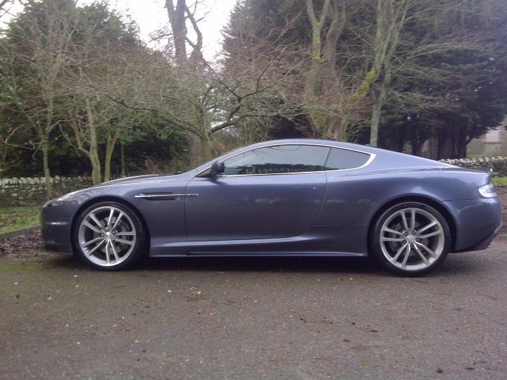 DBS wheels on DB9 - (more or less) - Page 1 - Aston Martin - PistonHeads - The image features a sleek, metallic blue Aston Martin GT4 coupe parked on an asphalt surface. The car is viewed from the rear three-quarter angle, highlighting the curvaceous design of the car, with its bulging fender and large alloy wheels. The Aston Martin emblem is clearly visible on the car's trunk lid, just behind the rear wheel. The background is a natural setting, with a variety of trees and underbrush, suggesting a serene, park-like environment. The sky appears overcast, casting diffused light on the car and the surroundings.