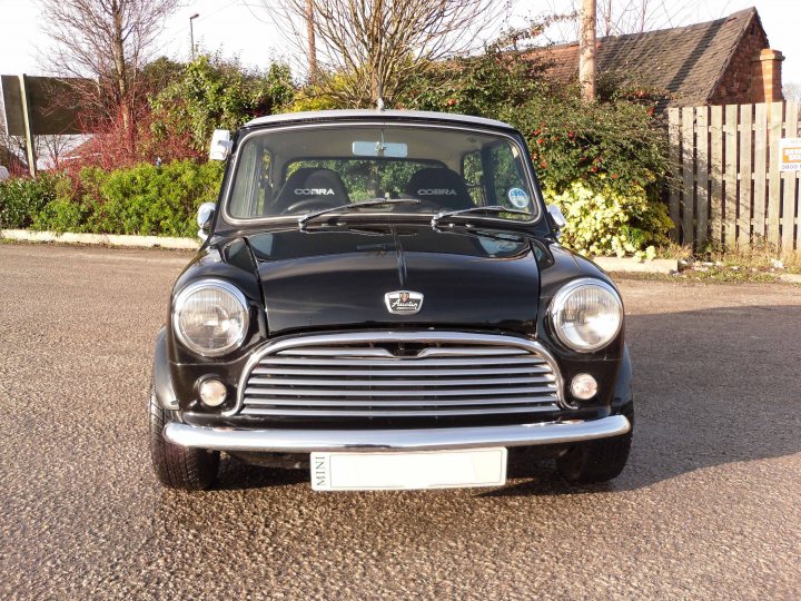 Sell or keep, MOT or don't? - Page 1 - Classic Minis - PistonHeads - The image shows a black mini Cooper car parked on an empty street next to a wooden fence. The car is positioned at an angle to the camera, allowing a clear view of its front and left side. The car is positioned on a paved street with trees and a hedge in the background. The car's license plate is clearly visible, displaying a foreign script and confirming the vehicle's registration details.