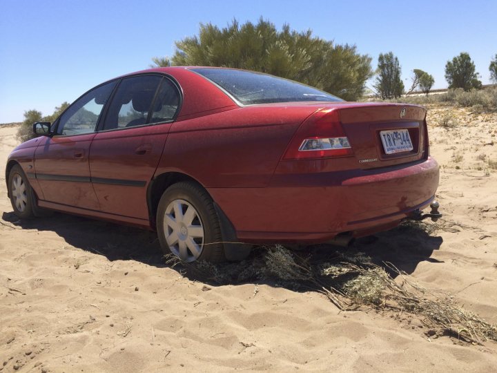 A yellow car is parked on the beach - Pistonheads - In the image, a vibrant red car is the main subject, as it stands out against the desert landscape. The car is stuck in the sand, tilted to the left, as if it has been racing on the sandy path before it came to a halt. The scene is set in a landscape of vast sand dunes, and the car's modest size compared to the sand dunes emphasizes its isolation in the environment. The sky is a brilliant blue, suggesting a clear and sunny day. The scene conveys the sense of adventure and the sheer scale of nature.