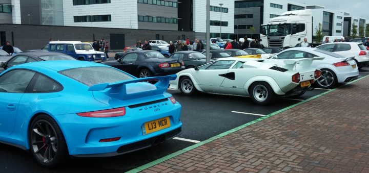 A car is parked on the side of the road - Pistonheads - The image shows a parking lot filled with various sports cars parked closely to each other. The cars have hoods that are opened wide or are hoodless, showcasing their engines. The ground appears wet, possibly due to recent rain, reflecting lights from the vehicles. In the background, there are tall buildings, indicating that the parking lot is in an urban environment. To the right, there's a large truck, which seems out of place among the sleek cars. Cars are parked on three sides, leaving no front or rear view for most of them.