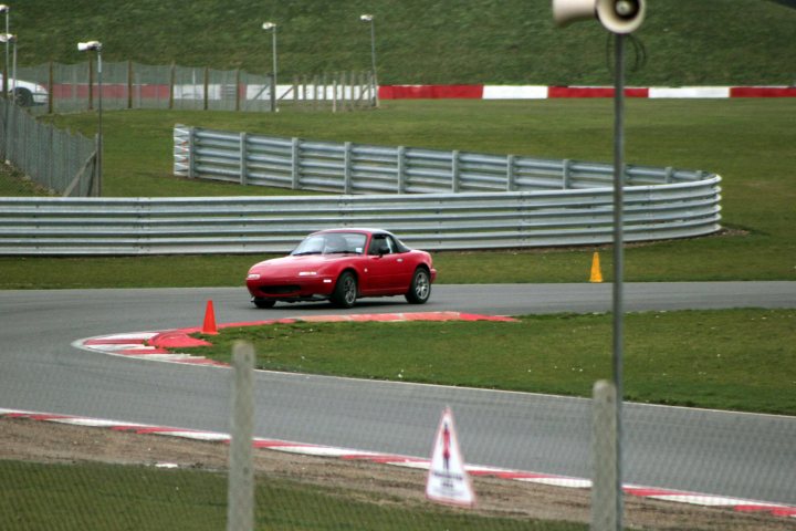 Track day at Snetterton Friday 15th February - Page 1 - East Anglia - PistonHeads - The image shows a red sports car driving along a track at high speed. The track appears to be a race track, with multiple camera rigs mounted on poles lining the tarmac. There are orange traffic cones and a red/white striped obstacle placed before a pole with a camera mounted on it. The setting is outdoors during the daytime, under bright, natural light. The car's speed and the presence of the cones suggest a training or practice scenario for a driver.