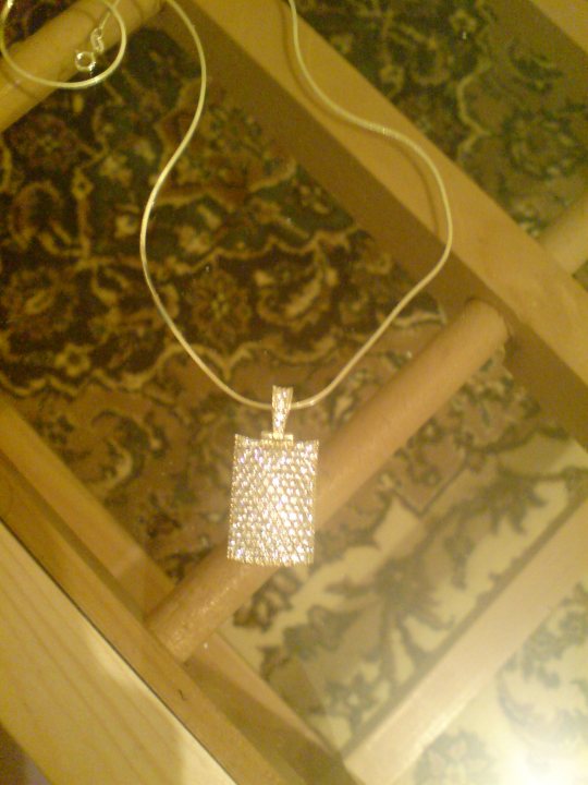 The image features a necklace with a sizable, diamond-studded pendant resting on a piece of darkly colored furniture. The pendant is a prominent, rectangular adornment. The necklace is suspended in mid-air, held by two white rings at the ends. The larger section of the furniture shows a dark, intricately designed pattern, while the smaller part displays a white, geometric pattern. The pendant is positioned closer to the viewer than the rest of the jewelry, suggesting it is the main focus of the image.