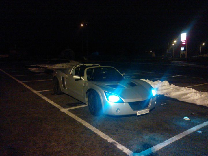 JFReturn's VX220 2.2 NA - Page 1 - VX - PistonHeads - The image features a modern sports car parked in the middle of a parking lot at night. The car is sleek and silver, exuding a sense of speed and luxury. It's positioned in the first parking spot, suggesting that someone has just arrived or is about to depart. The lot is spacious and mostly empty, indicating low activity or visitation here. The lighting is artificial and enhances the hard lines and angles of the car's design. The darkness around further emphasizes the car's striking appearance.