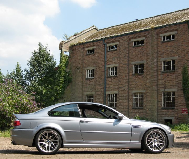 Local photo locations - Page 1 - East Anglia - PistonHeads - The image depicts a gray BMW sports car parked on a gravel surface in front of a large brick building. The building appears to be a historical structure, with several arched windows filled with greenery, suggesting a connection with nature. The sky is clear with a few clouds, and there's a touch of moss or ivy on the upper part of the barn, blending the constructed space with nature. It seems to be held on a sunny day.