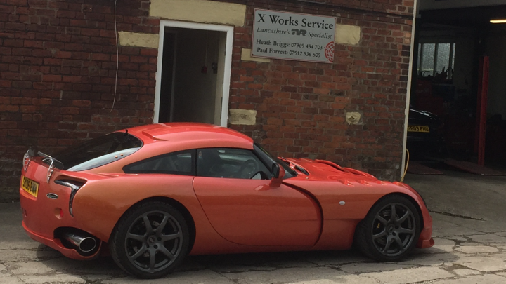 Tamora 4.3 1st 12k service and visit to X Works Nr Kirkham - Page 1 - Tamora, T350 & Sagaris - PistonHeads - The image features a vibrant orange sports car parked outside a building. The car's top is down, revealing a luxurious black interior. The vehicle is parked on the street, and a yellow license plate is clearly visible on the rear. The brick building in the background has a simple sign that reads "X WORKS SERVICE", indicating the business nature of the establishment.