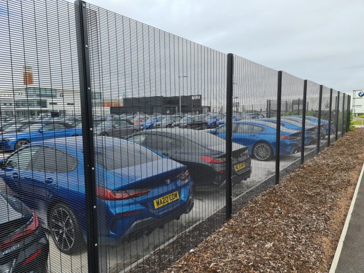 840i M Sport Lease Deal - Page 138 - BMW General - PistonHeads - The image shows a parking lot filled with numerous cars, predominantly blue and black in color. A black wire fence encloses the area, separating it from another space. Behind the fence, there's a grassy area and trees, suggesting this might be a public or communal parking area rather than a private one. The cars are neatly parked, with some showing their license plates, indicating that they are personal vehicles. There's no visible activity in the lot at the moment of the photo.