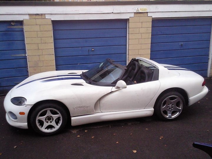 Pistonheads Viper Rebuild - The image shows a white sports car parked against a backdrop of blue garage doors. The vehicle features a distinctive blue stripe on the hood and a black convertible top. The car is positioned with the door and rear windshield open, revealing what appears to be a single black seat visible through the window. The setting suggests a residential area, possibly the driveway of a home.