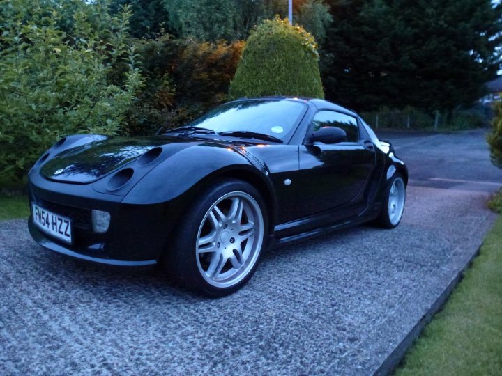 Pistonheads - The image features a black Mazda Miata sports car parked on a gray, asphalt driveway. The vehicle is raked at a sharp angle which gives a sense of its dynamic potential. It's a convertible car with the door on the driver side. The setting is residential with healthy green bushes present on both sides of the image, providing a natural border to the scene.